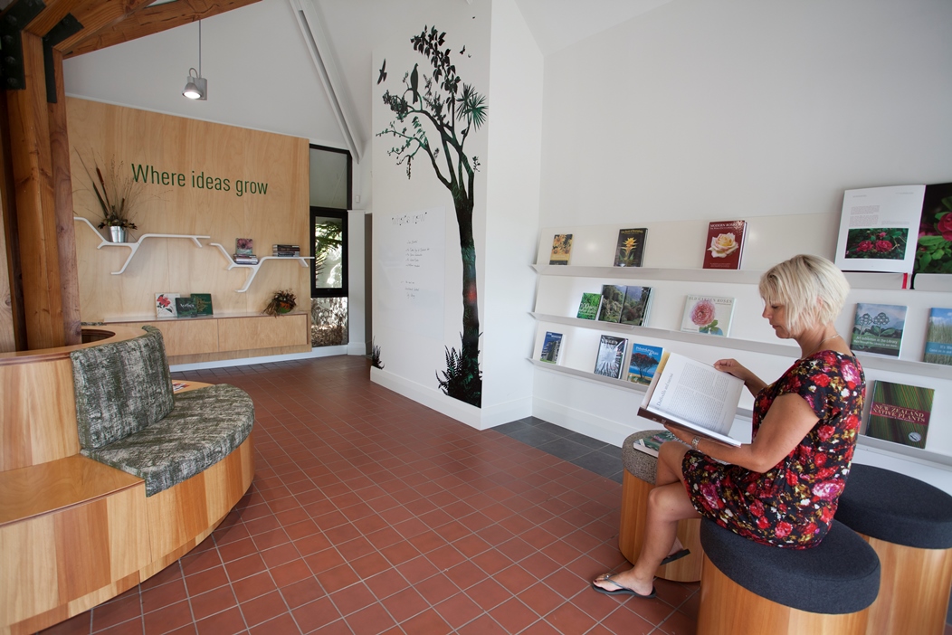 A woman in floral dress sits reading a book on a comfy seat in the foyer of the Auckland Botanic Gardens library.