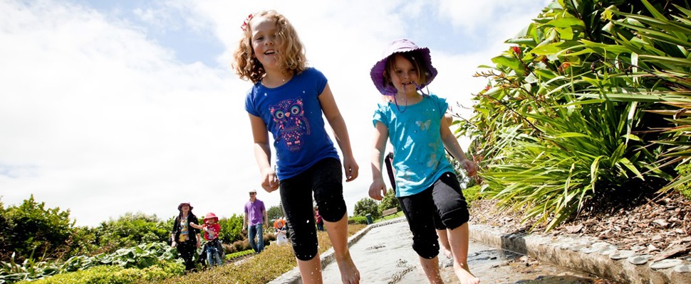 Taken from low down the photo shows two children running barefoot through a channel of water on a warm sunny day at Auckland Botanic Gardens