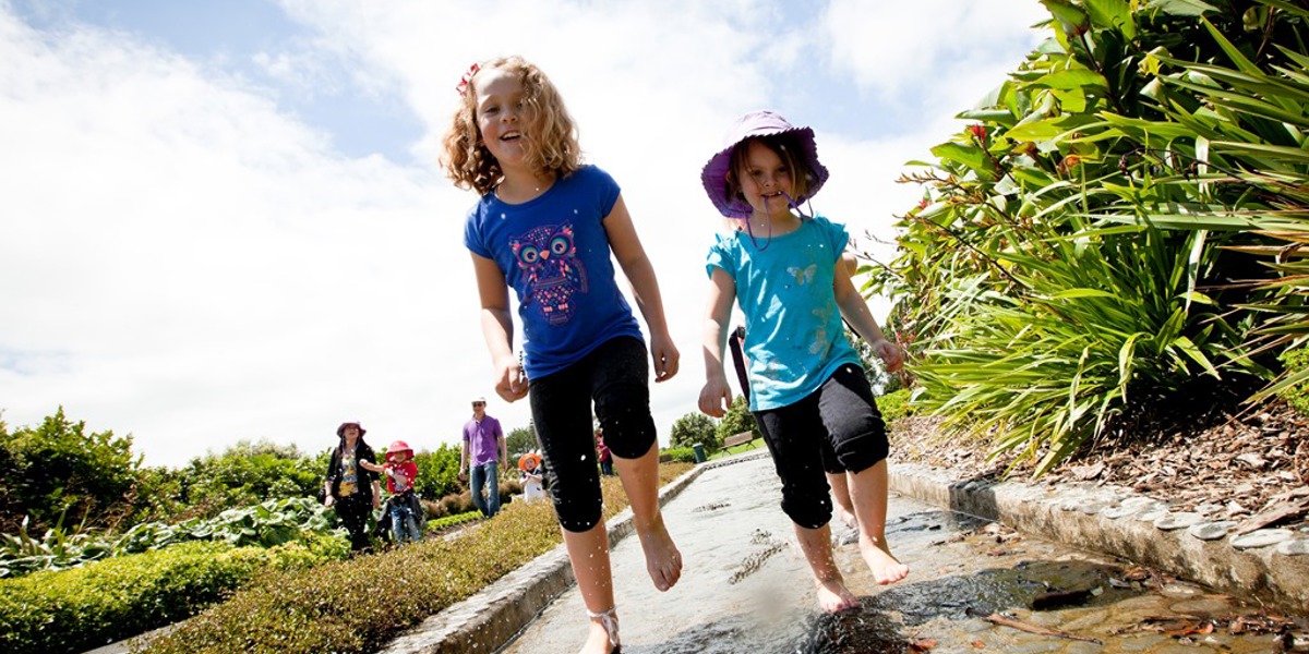 Taken from low down the photo shows two children running barefoot through a channel of water on a warm sunny day at Auckland Botanic Gardens