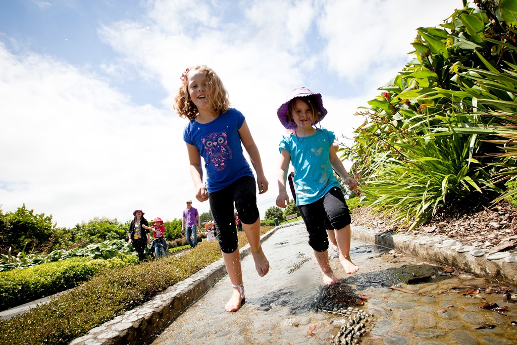 Taken from low down the photo shows two children running barefoot through a channel of water on a warm sunny day at Auckland Botanic Gardens
