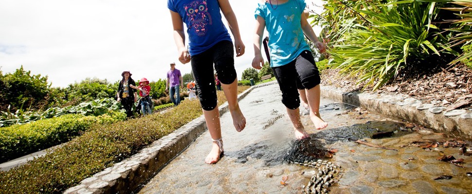 Taken from low down the photo shows two children running barefoot through a channel of water on a warm sunny day at Auckland Botanic Gardens