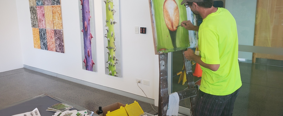 Artist Neal Palmer is working at his easel. He has his back to the camera and paintbrush in hand. He is applying paint to a work in progress that will be a close up of a thorn. In the background are some of Neal's finished works of bark rubbings and large scale plant stems covered in thorns. In the foreground are an apparently chaotic arrangement of papers and photographs Neal has taken of various plants during his time as artist in residence at Auckland Botanic Gardens.